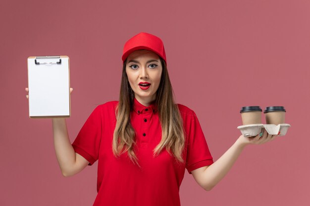 Mensajero femenino de vista frontal en uniforme rojo sosteniendo tazas de café de entrega y bloc de notas en el uniforme de trabajador de trabajo de entrega de servicio de fondo rosa