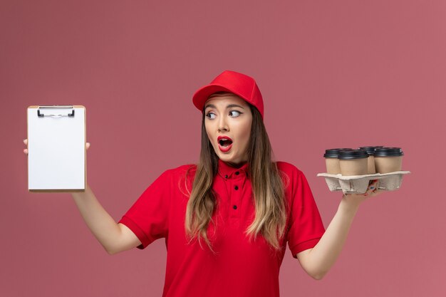 Mensajero femenino de vista frontal en uniforme rojo sosteniendo tazas de café de entrega y bloc de notas en uniforme de trabajo de empresa de entrega de servicio de fondo rosa claro