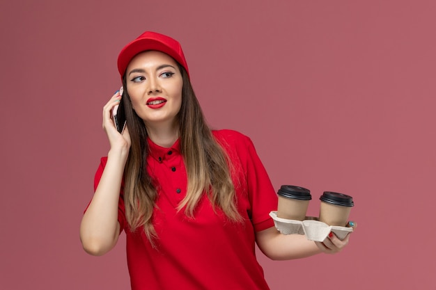 Mensajero femenino de vista frontal en uniforme rojo sosteniendo tazas de café de entrega hablando por teléfono en el uniforme de entrega de servicio de fondo rosa