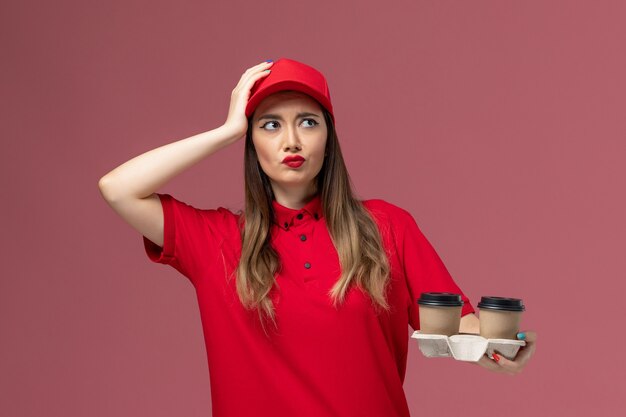 Mensajero femenino de vista frontal en uniforme rojo sosteniendo tazas de café de entrega marrón y pensando en fondo rosa claro servicio entrega uniforme trabajador trabajo empresa femenina