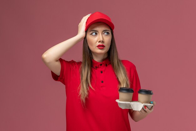 Mensajero femenino de vista frontal en uniforme rojo sosteniendo tazas de café de entrega marrón pensando en uniforme de entrega de servicio de fondo rosa claro