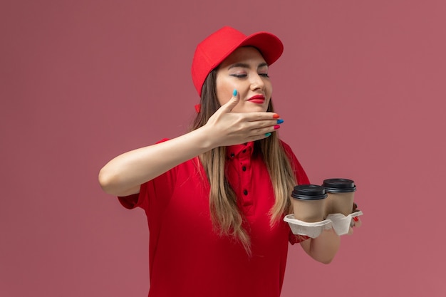 Mensajero femenino de vista frontal en uniforme rojo sosteniendo tazas de café de entrega marrón que huelen a uniforme de entrega de servicio de fondo rosa claro