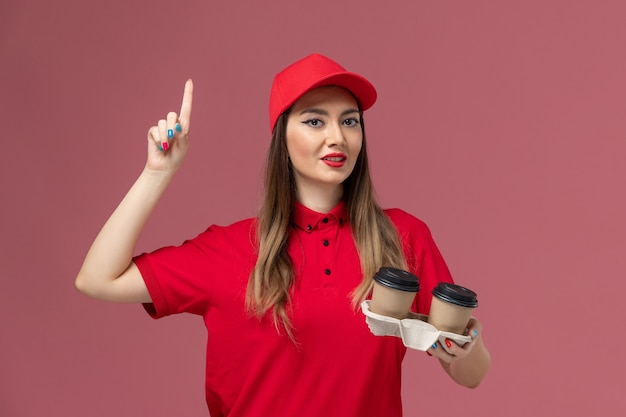 Mensajero femenino de vista frontal en uniforme rojo sosteniendo tazas de café de entrega marrón sobre fondo rosa claro servicio de entrega uniforme empresa de trabajadores