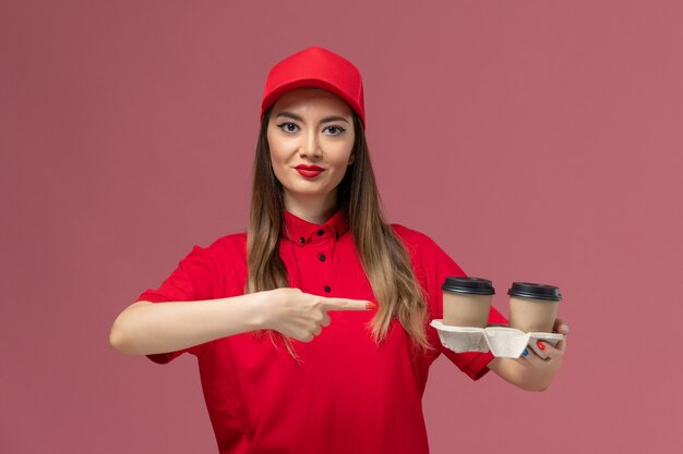 Mensajero femenino de vista frontal en uniforme rojo sosteniendo tazas de café de entrega marrón sobre fondo rosa claro servicio entrega uniforme trabajador trabajo mujer dama empresa