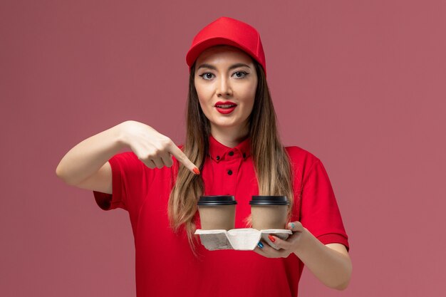 Mensajero femenino de vista frontal en uniforme rojo sosteniendo tazas de café de entrega marrón en el trabajo uniforme de entrega de servicio de fondo rosa claro