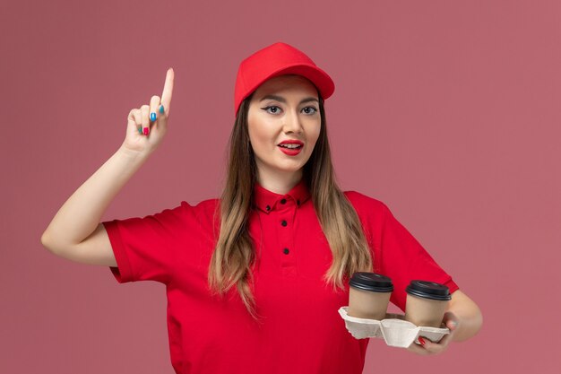Mensajero femenino de vista frontal en uniforme rojo sosteniendo tazas de café de entrega marrón en el trabajo uniforme de entrega de servicio de fondo rosa