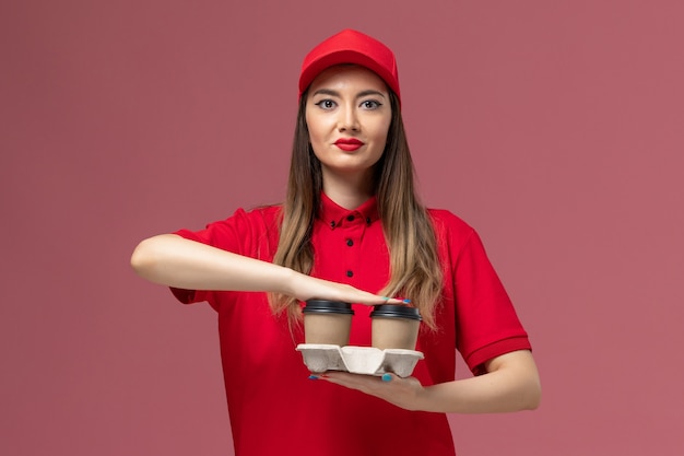 Mensajero femenino de vista frontal en uniforme rojo sosteniendo tazas de café de entrega marrón en el uniforme de entrega de trabajo de servicio de fondo rosa claro
