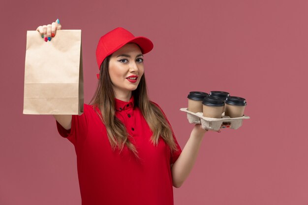 Mensajero femenino de vista frontal en uniforme rojo sosteniendo tazas de café de entrega con paquete de alimentos con una leve sonrisa en uniforme de trabajo de entrega de servicio de fondo rosa