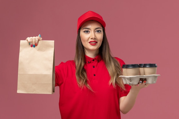 Mensajero femenino de vista frontal en uniforme rojo sosteniendo tazas de café de entrega con paquete de alimentos en el uniforme de trabajo de entrega de servicio de fondo rosa