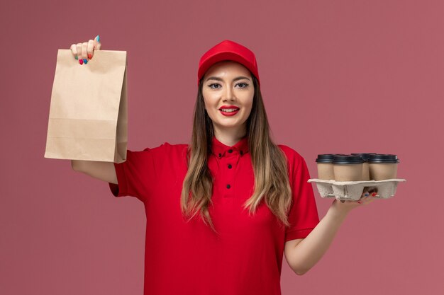 Mensajero femenino de vista frontal en uniforme rojo sosteniendo tazas de café de entrega con paquete de comida en el uniforme de trabajo de entrega de servicio de escritorio rosa