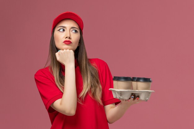 Mensajero femenino de vista frontal en uniforme rojo sosteniendo tazas de café de entrega pensando en el trabajador uniforme de trabajo de entrega de servicio de fondo rosa