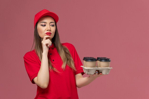 Mensajero femenino de vista frontal en uniforme rojo sosteniendo tazas de café de entrega pensando en el trabajador uniforme de trabajo de entrega de servicio de fondo rosa