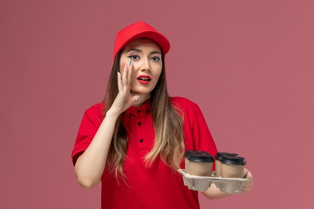 Mensajero femenino de vista frontal en uniforme rojo sosteniendo tazas de café de entrega susurrando sobre fondo rosa trabajador de trabajo uniforme de entrega de servicio