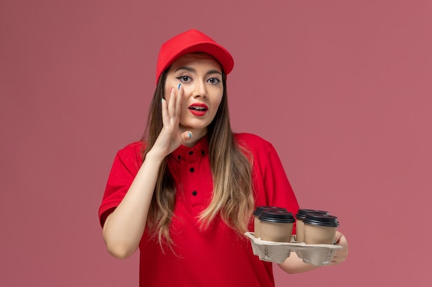 Mensajero femenino de vista frontal en uniforme rojo sosteniendo tazas de café de entrega susurrando sobre fondo rosa trabajador de trabajo uniforme de entrega de servicio