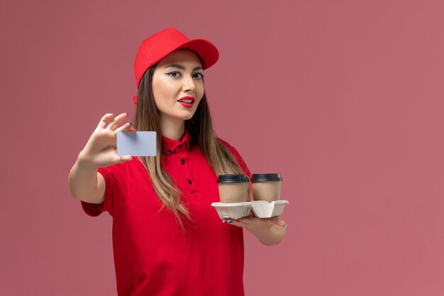 Mensajero femenino de vista frontal en uniforme rojo sosteniendo tazas de café de entrega y tarjeta posando sobre fondo rosa uniforme de entrega de trabajo de servicio