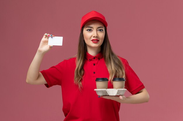 Mensajero femenino de vista frontal en uniforme rojo sosteniendo tazas de café de entrega y tarjeta en el uniforme de entrega de trabajo de servicio de fondo rosa