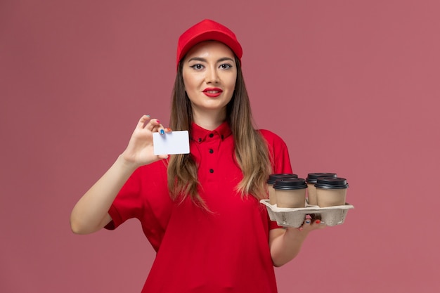 Mensajero femenino de vista frontal en uniforme rojo sosteniendo tazas de café de entrega y tarjeta en uniforme de trabajo de entrega de servicio de fondo rosa