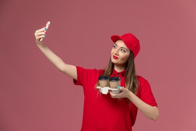 Mensajero femenino de vista frontal en uniforme rojo sosteniendo tazas de café de entrega tomando fotos con ellos en uniforme de entrega de servicio de trabajo de trabajador de fondo rosa