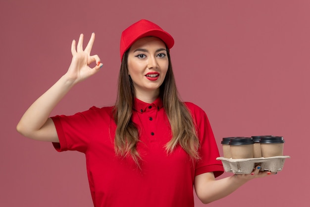 Mensajero femenino de vista frontal en uniforme rojo sosteniendo tazas de café de entrega en el trabajador de trabajo uniforme de entrega de servicio de escritorio rosa