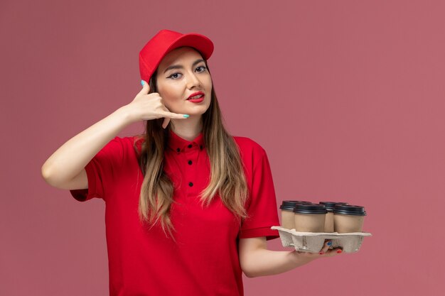 Mensajero femenino de vista frontal en uniforme rojo sosteniendo tazas de café de entrega en el trabajador uniforme de trabajo de entrega de servicio de fondo rosa