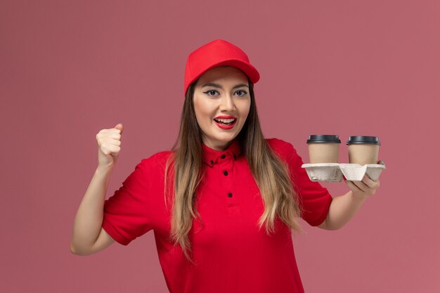 Mensajero femenino de vista frontal en uniforme rojo sosteniendo tazas de café de entrega en el uniforme de entrega de servicio de trabajo de trabajador de fondo rosa