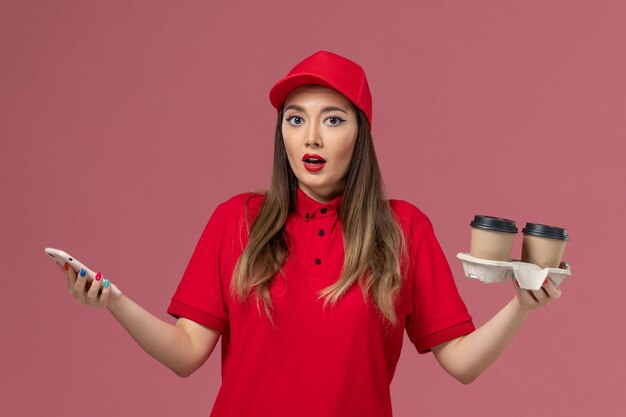 Mensajero femenino de vista frontal en uniforme rojo sosteniendo tazas de café de entrega y usando el teléfono en uniforme de entrega de servicio de trabajo de trabajador de fondo rosa claro