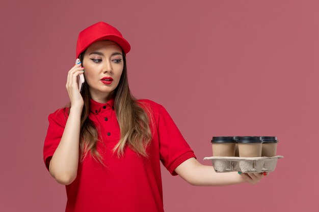 Mensajero femenino de vista frontal en uniforme rojo sosteniendo tazas de café y hablando por teléfono en uniforme de entrega de servicio de escritorio rosa