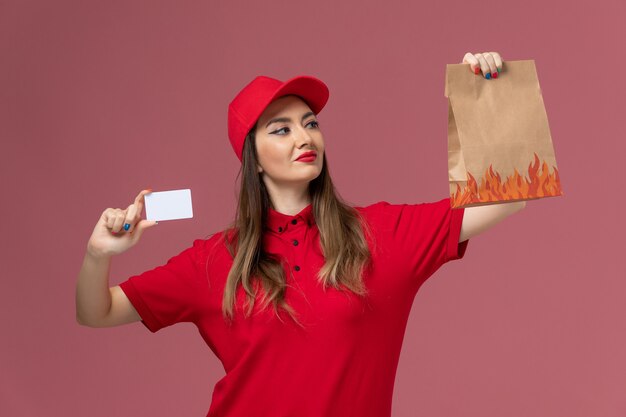 Mensajero femenino de vista frontal en uniforme rojo con tarjeta blanca y paquete de alimentos en la empresa de uniforme de entrega de servicio de fondo rosa