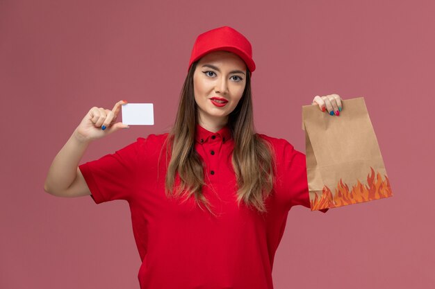 Mensajero femenino de vista frontal en uniforme rojo con tarjeta blanca y paquete de alimentos en la empresa de uniforme de entrega de trabajo de servicio de fondo rosa