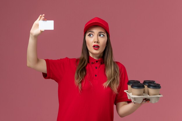 Mensajero femenino de vista frontal en uniforme rojo con tarjeta blanca sobre fondo rosa claro servicio de entrega de trabajo empresa uniforme