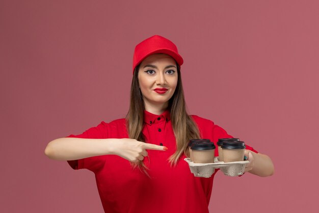 Mensajero femenino de vista frontal en uniforme rojo con tazas de café de entrega posando sonriendo en el trabajo uniforme de entrega de servicio de fondo rosa