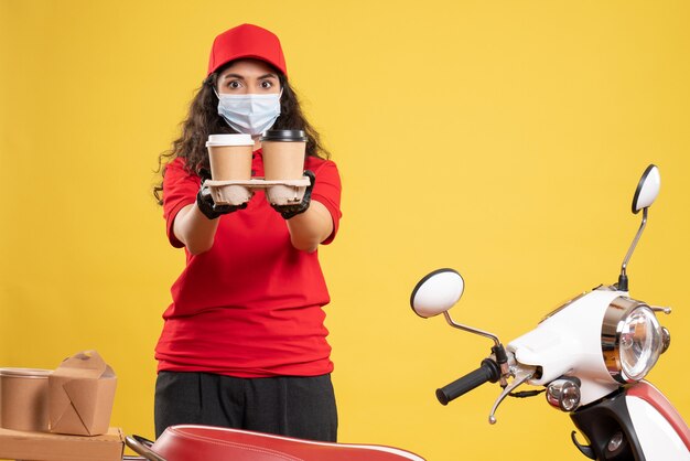 Mensajero femenino de vista frontal en uniforme rojo con tazas de café sobre fondo amarillo entrega del trabajador covid- virus uniforme de trabajo pandémico
