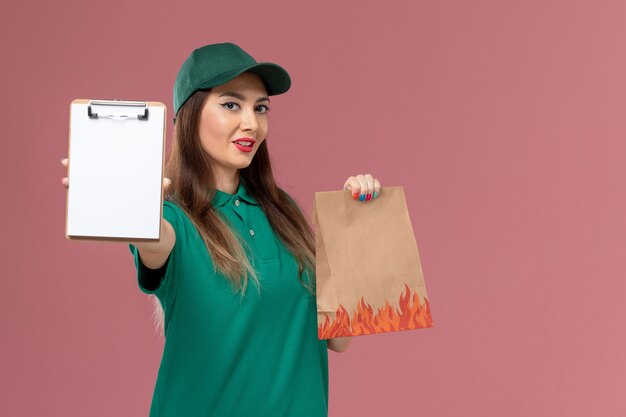 Mensajero femenino de vista frontal en uniforme verde con bloc de notas y paquete de alimentos en el trabajador de trabajo de entrega uniforme de servicio de pared rosa