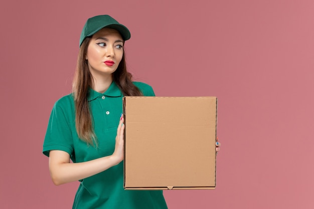 Mensajero femenino de vista frontal en uniforme verde con caja de comida en la pared de color rosa claro servicio de trabajo uniforme de entrega