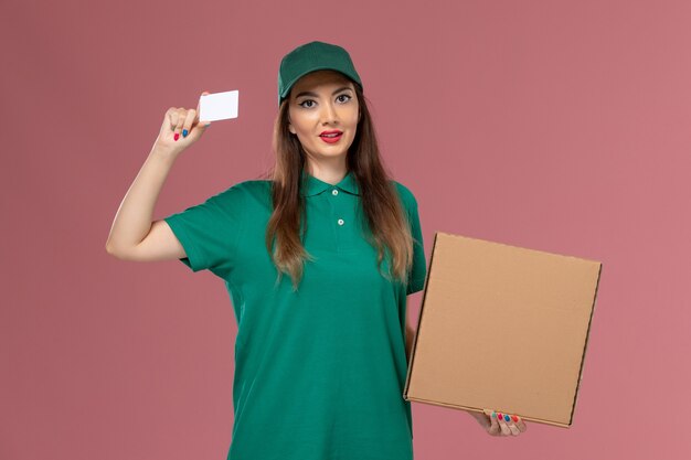 Mensajero femenino de vista frontal en uniforme verde con caja de entrega de alimentos y tarjeta en la pared rosa claro trabajador de trabajo de entrega uniforme de servicio de empresa