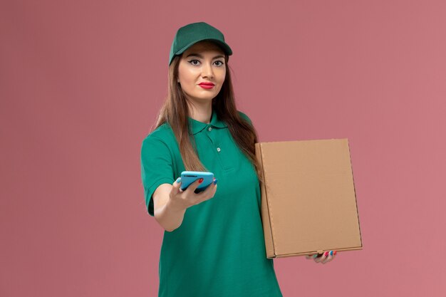 Mensajero femenino de vista frontal en uniforme verde con caja de entrega de alimentos usando el teléfono en el trabajo de entrega uniforme de servicio de pared rosa