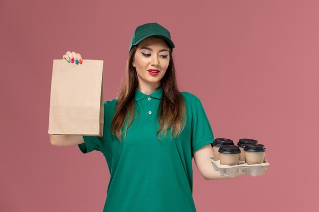 Mensajero femenino de vista frontal en uniforme verde y capa con paquete de alimentos y tazas de café de entrega en el trabajo de entrega uniforme de servicio de escritorio rosa