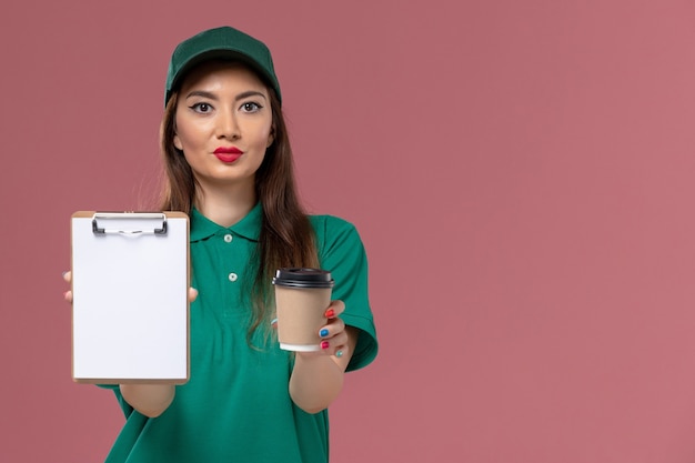 Mensajero femenino de la vista frontal en uniforme verde y capa que sostiene la taza de café de la entrega y el bloc de notas en la empresa femenina de la entrega uniforme del trabajo del servicio de la pared rosa