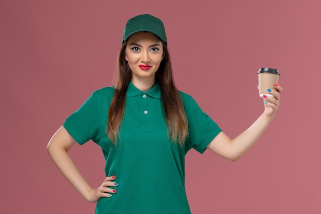 Mensajero femenino de la vista frontal en uniforme verde y capa que sostiene la taza de café de la entrega en la entrega uniforme del trabajo del servicio de la pared rosa clara