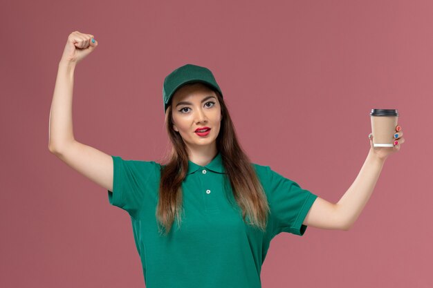 Mensajero femenino de vista frontal en uniforme verde y capa sosteniendo la taza de café de entrega animando en el trabajo de entrega uniforme de trabajo de servicio de pared rosa