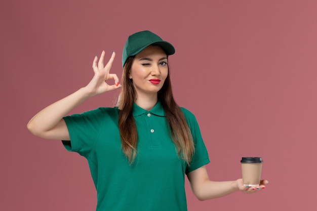 Foto gratuita mensajero femenino de vista frontal en uniforme verde y capa sosteniendo la taza de café de entrega en la pared rosa trabajo de servicio uniforme de trabajo de trabajador de entrega