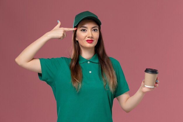 Mensajero femenino de vista frontal en uniforme verde y capa sosteniendo la taza de café de entrega sonriendo en el trabajo de entrega uniforme de servicio de pared rosa