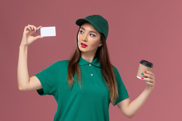 Mensajero femenino de vista frontal en uniforme verde y capa sosteniendo la taza de café de entrega con tarjeta en el trabajo de entrega uniforme de servicio de pared rosa