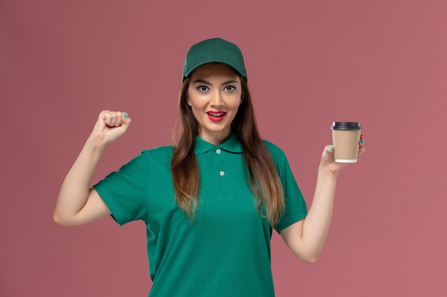 Mensajero femenino de vista frontal en uniforme verde y capa sosteniendo la taza de café de entrega en el trabajador de entrega uniforme de trabajo de servicio de pared rosa