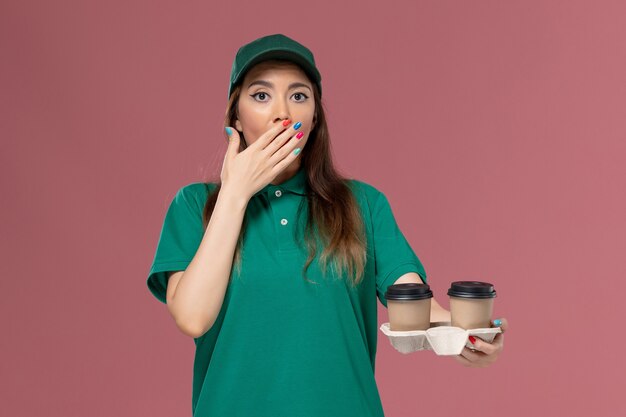 Mensajero femenino de vista frontal en uniforme verde y capa sosteniendo tazas de café de entrega en la pared rosa claro servicio de la empresa trabajo entrega uniforme