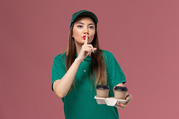 Mensajero femenino de vista frontal en uniforme verde y capa sosteniendo tazas de café de entrega en la pared rosa servicio de empresa entrega uniforme trabajo femenino niña