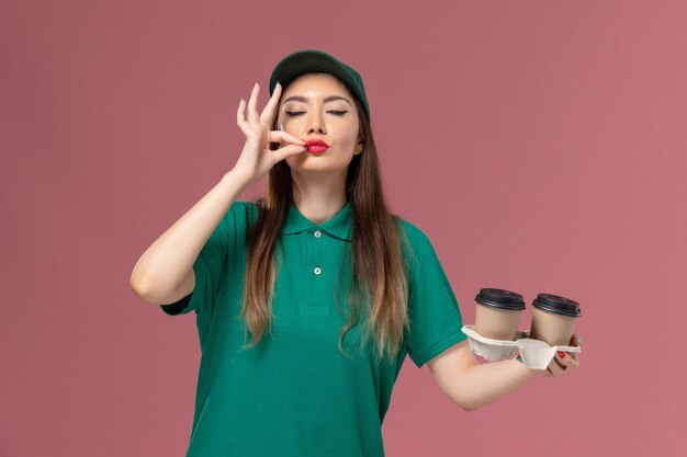 Mensajero femenino de vista frontal en uniforme verde y capa sosteniendo tazas de café de entrega en la pared rosa servicio de la empresa uniforme entrega trabajadora trabajo femenino