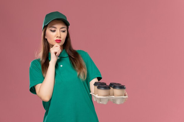 Mensajero femenino de vista frontal en uniforme verde y capa sosteniendo tazas de café de entrega y pensando en el trabajo de entrega uniforme de servicio de pared rosa