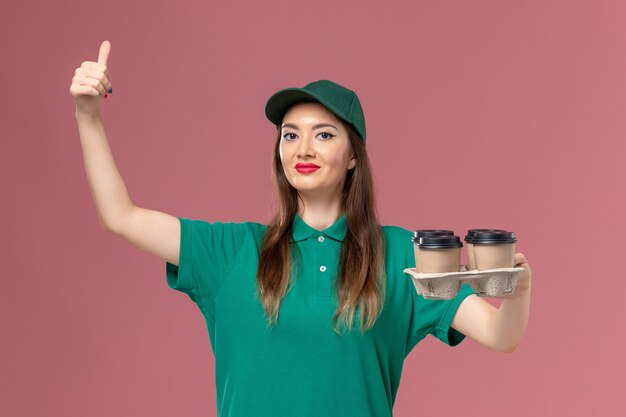 Mensajero femenino de vista frontal en uniforme verde y capa sosteniendo tazas de café de entrega posando en servicio de pared rosa trabajador de trabajo de entrega uniforme