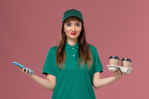 Mensajero femenino de vista frontal en uniforme verde y capa sosteniendo tazas de café de entrega y su teléfono en servicio de escritorio rosa entrega uniforme de trabajo